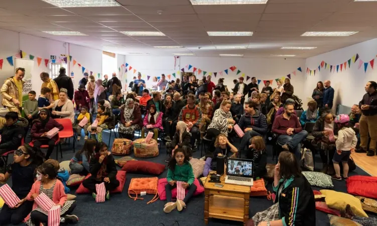 Room with colourful bunting around the walls. Approximately 50 people of all ages sit on floor cushions and chairs.