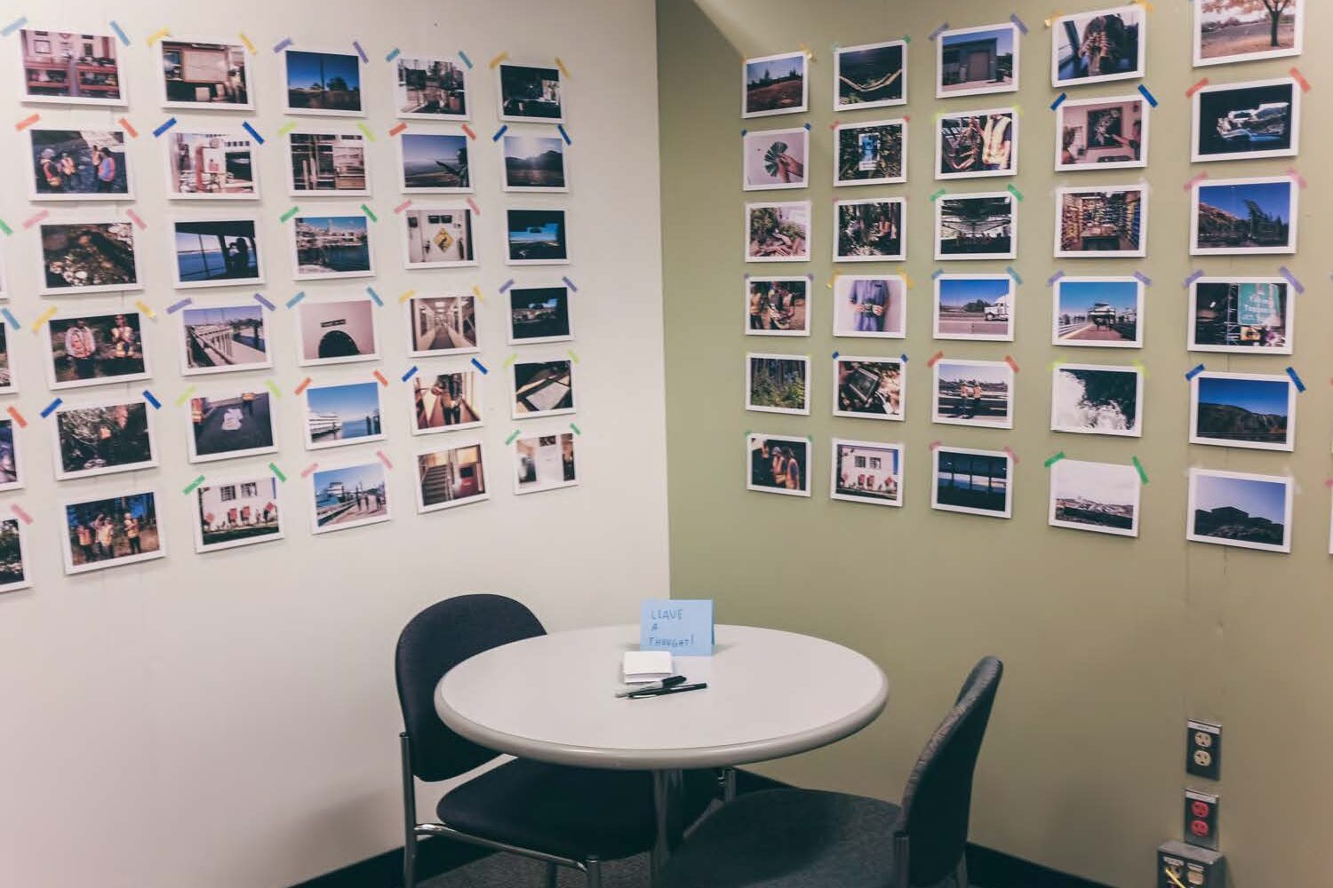 An office space with a small table and two chairs. Behind, small photographs are taped to the walls.