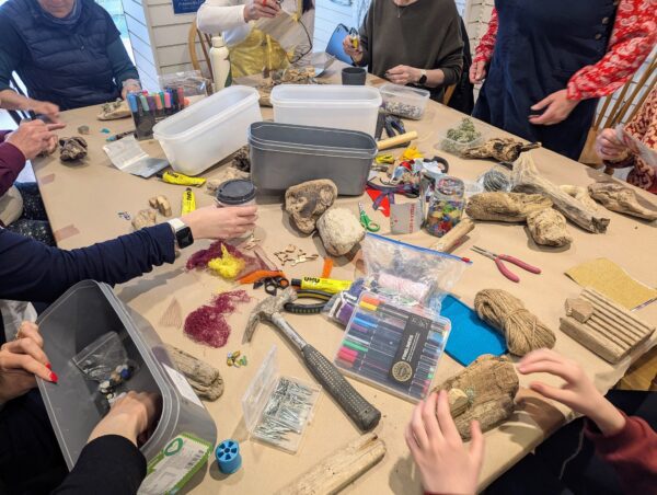 A photo of a messy table with a mixture of stones, natural materials and craft materials. People's hands are reaching out to take objects to make things with.