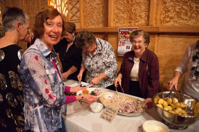 Women serving food from a buffet table. Two women look into the camera, both laughing.