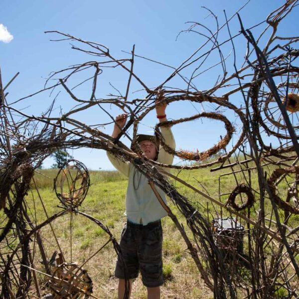 Young person reaches above their head to weave a large wicker sculpture.