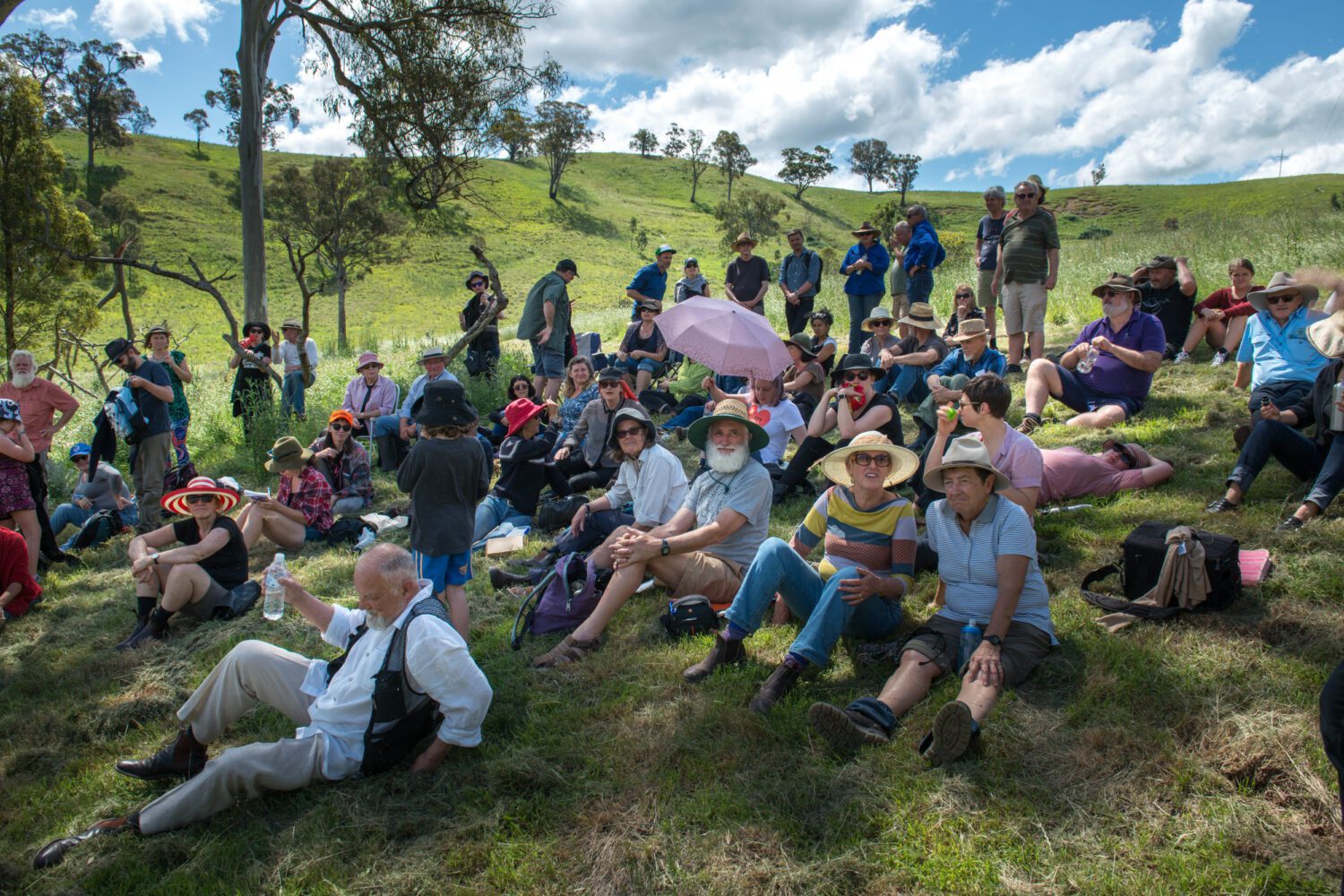 Group of 40+ people sit on grass, in the shade of a tree. Valley setting.