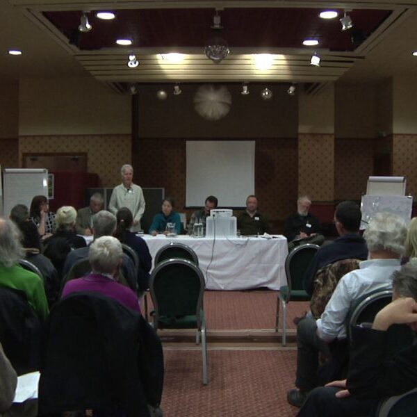 People sitting in a small auditorium. The stage has a panel of six people, and there are rows of seats facing it.