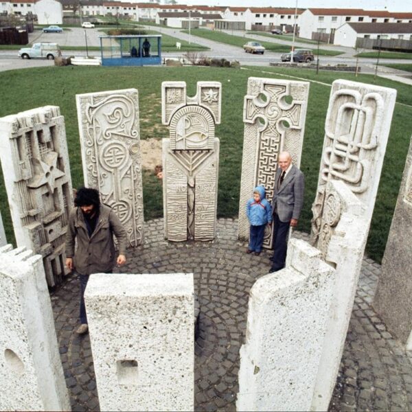 Two adults and a child stand in the centre of a stone circle, comprised of 13 concrete slabs. In the background is a housing estate.