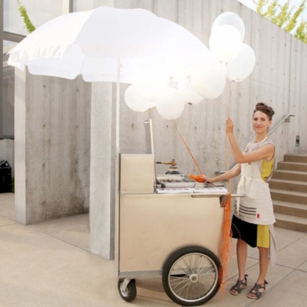 Artist next to a cart with a large umbrella, holding white balloons.