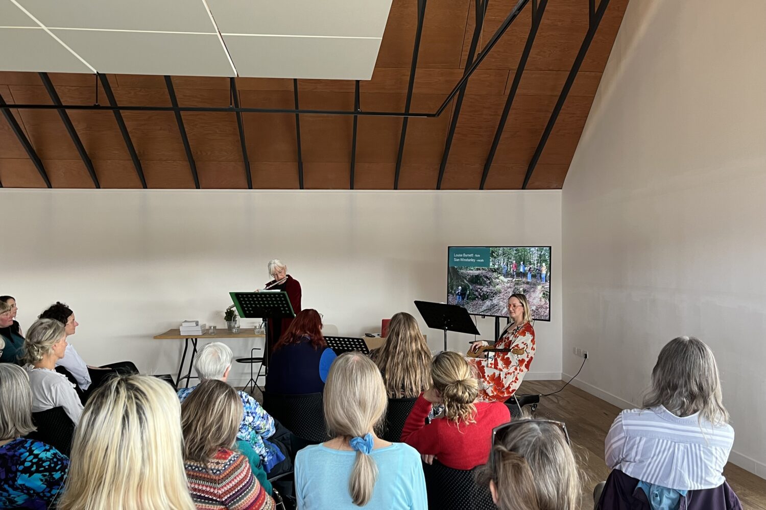 A group of people are seating, listening to a woman play the flute at the front of the room, while another woman plays ukulele.