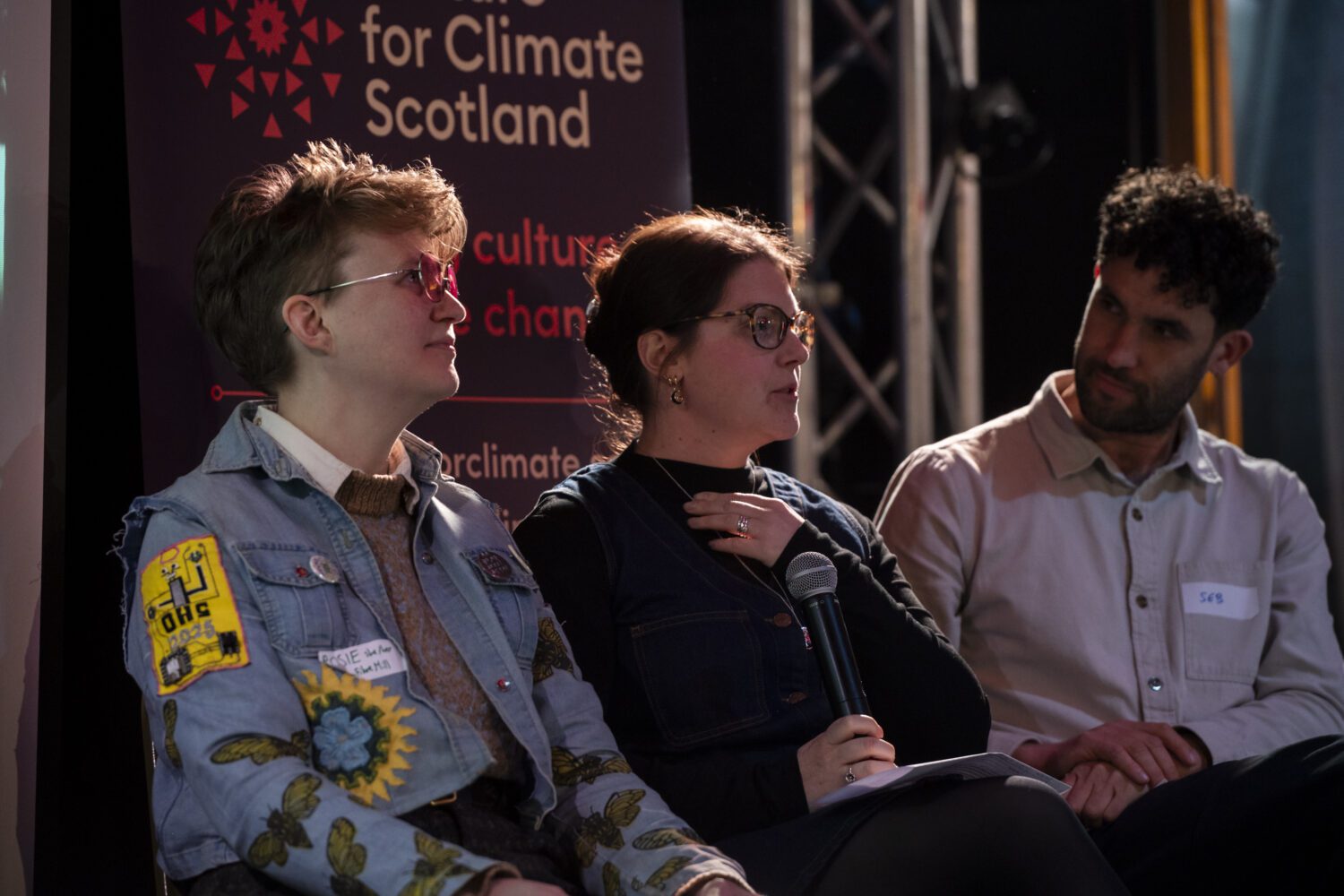 Three people sat down. The woman in the middle holds a microphone and is talking.