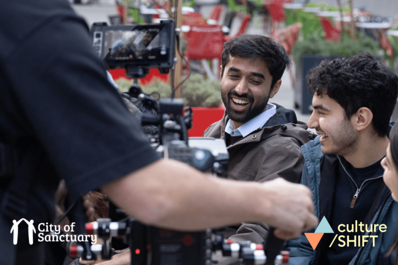 Two men laugh while being filmed by a professional camera. Logos for Culture for Climate Scotland and City of Sanctuary UK