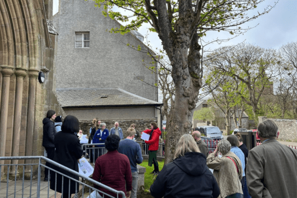A group of people gathered outside a stone building with an arched entrance. Some hold papers, suggesting a guided tour. Trees with budding leaves surround them.